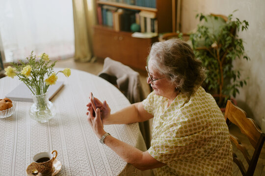 Senior Caucasian woman sitting at round table using smartphone, smiling while interacting with device, vase with yellow flowers and cup of coffee placed on table nearby