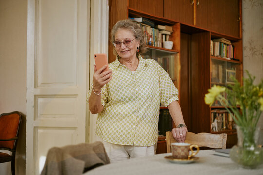 Senior Caucasian woman standing and smiling while holding smartphone, engaging in video call or checking messages in home setting, bookshelf and flowers visible in background