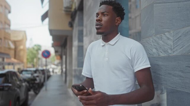 Man in white polo shirt holds smartphone and glances upward along stone building on busy city street; contemplation.