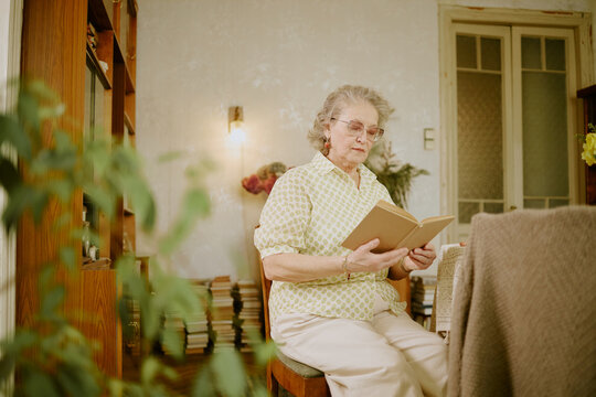 Senior Caucasian woman sitting indoors reading book, wearing eyeglasses, appearing focused and engaged with literature, surrounded by bookshelves and home decor elements