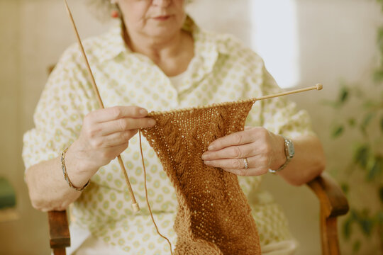 Senior Caucasian woman sitting and knitting with wooden needles, holding partially completed brown scarf, focusing on intricate handwork, wearing patterned shirt, indoors