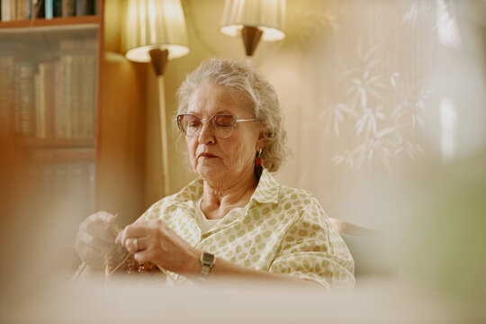Senior Caucasian woman sitting indoors knitting with focused expression, wearing glasses and patterned shirt, hands holding yarn, surrounded by warm lighting and blurred background elements
