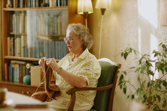 Senior Caucasian woman sitting in armchair knitting with yarn, wearing eyeglasses, focusing on craft project in cozy living room with bookshelves and houseplant in background