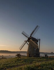 Naklejka premium A traditional windmill stands serenely in a rural landscape at sunrise, its stone façade reflecting warm golden light amidst a clear blue sky.