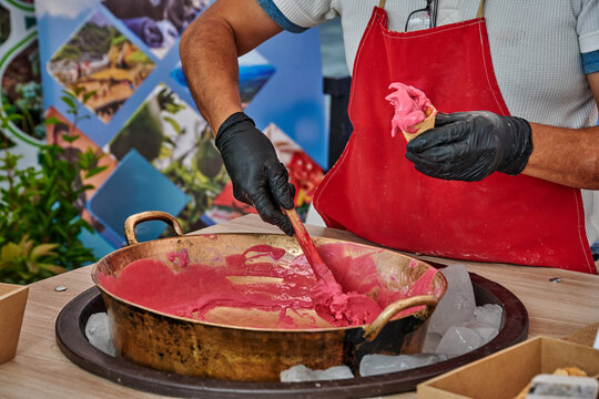 Ecuadorian chef prepares traditional dessert helado de paila ice cream in large metal bowl on pieces of ice, wearing gloves and an apron, with colorful background elements and ice visible on table