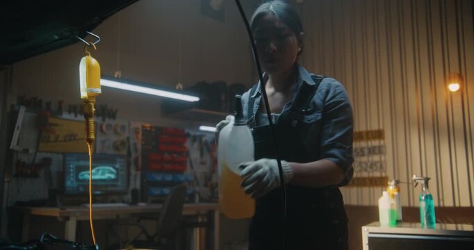 Skilled Asian Woman in Overalls Performs Car Maintenance Pouring Liquid Into Engine Compartment. Lined Up By Tools, Girl Demonstrates Precision, Responsibility, and Modern Mechanical Professionalism.