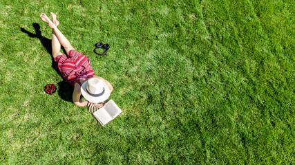 Young woman reading book in park in summer, girl relaxing outdoors on grass with book and headphones, analog lifestyle and digital detox trend, aerial drone view from above