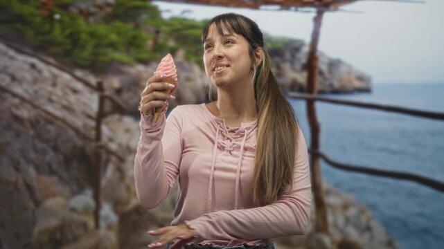 Young woman holding a pink softserve ice cream cone, hand on hip and smiling while looking up on a cliffside forest; carefree joy.