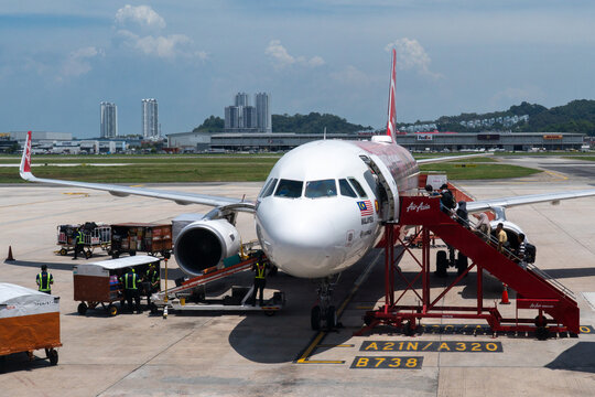 A front view of an AirAsia Airbus A320 aircraft at an airport gate. Passengers are boarding via a mobile staircase while ground crew and baggage handlers work around the left engine on a sunny day.