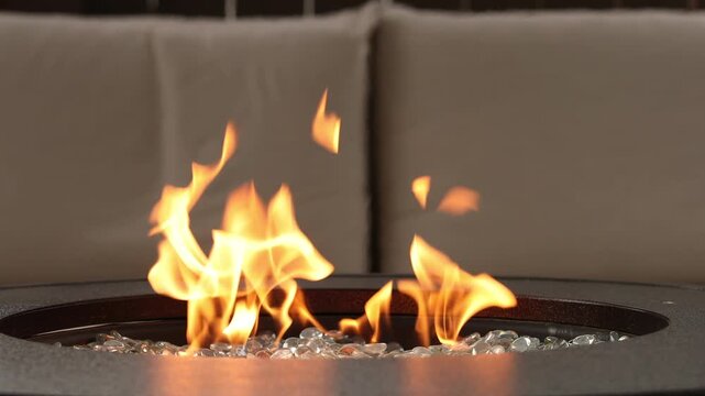 Modern outdoor gas fire pit with dancing yellow flames over glass beads and comfortable patio furniture background