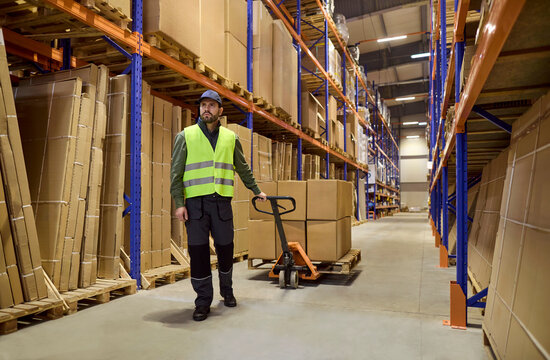 Worker in warehouse pulls pallet with jack. Large boxes on trolley move between shelves, storage logistics, shipping, and delivery. Lean warehouse operations and strong supply chain concept.