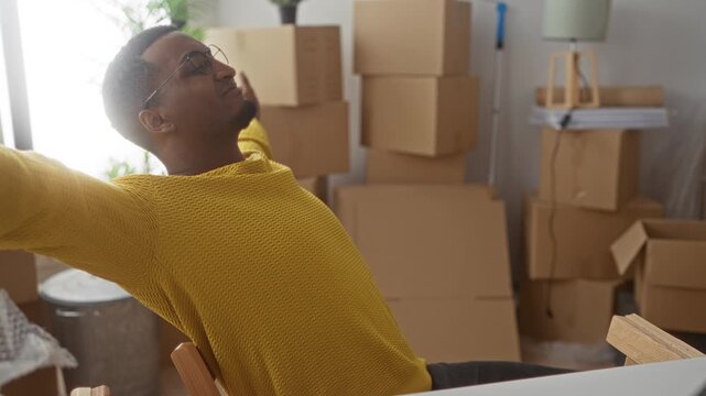 Man in yellow sweater and glasses with hands behind head stretching while seated amid stacked moving boxes in building; relief pause.