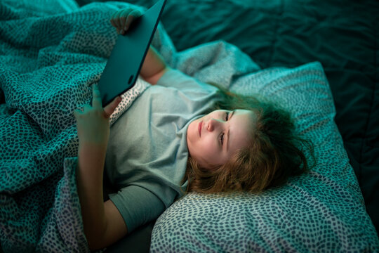 Teenager distracted by tablet screen in bed at night. Child spending night time with tablet in bed at night. Kid watching digital content on tablet in bed at night.