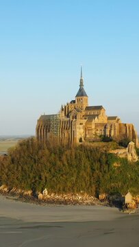 Scenic view of the historic Mont-Saint-Michel abbey bathed in warm morning light, surrounded by tidal sands and lush greenery in Normandy, France.