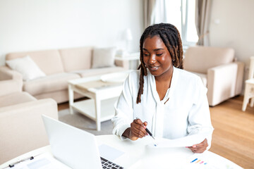 Smiling black woman reviewing documents at home office. Successful female freelancer holding papers...