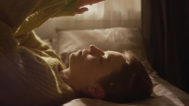 Close up shot of young adult woman lying down on bed touching hair while coping with anxious thoughts in sunlit bedroom