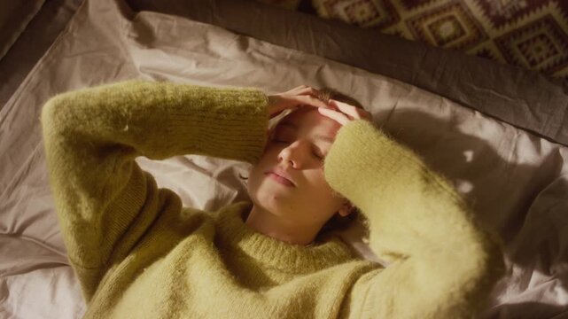 High angle shot of stressed young adult woman lying down on bed fidgeting with ring on finger then holding head feeling frustrated in sunlit bedroom