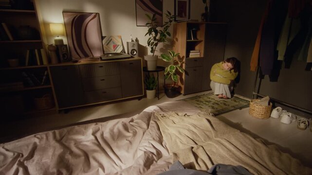 Wide angle shot of young adult woman sitting on bedroom floor clutching knees in sunlit room while going through painful heartbreak