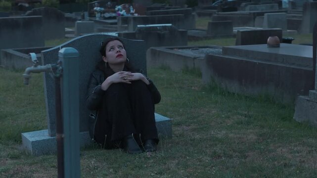 A young woman sits somberly by a tombstone at a Melbourne cemetery during sunset, embracing grief and reflection. The serene graveyard setting and soft lighting evoke contemplation.