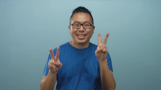 Young chinese man wearing blue t shirt and glasses smiling while flashing two peace signs with hands in studio against light blue wall; joy playful energy.