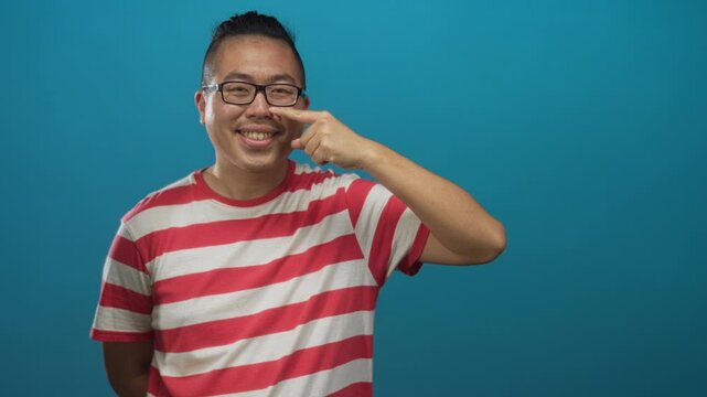Young chinese man points finger to nose in teal studio while smiling, wearing glasses and a red and white striped shirt, relaxed casual pose; playfulness.