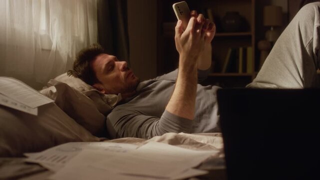 Rack focus of adult man checking smartphone struggling to get down to work on laptop while lying on bed cluttered with paperwork in low key lit bedroom