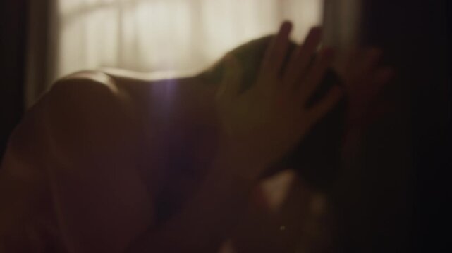 Handheld shot of shirtless adult man having outburst of anger throwing pillows trying to calm down while sitting on bed in backlight, shaky camera