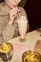 Vertical midsection shot of young woman drinking milkshake with whipped cream and sprinkles through straw while sitting at table with French fries in foreground