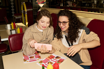 Fototapeta premium Caucasian young woman and Caucasian young man sitting at table in diner smiling and looking at smartphone together, holding menus with colorful food images, ketchup and mustard bottles visible
