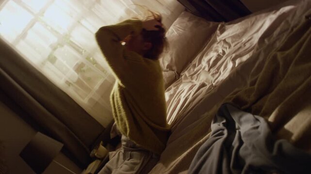 Side view shot of overwhelmed young adult woman holding head going through nervous breakdown then falling onto bed against window in sunlit room, roll camera movement