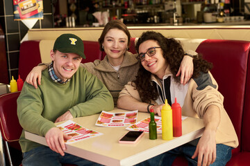 Fototapeta premium Portrait of three young adults, two Caucasian men and one Caucasian woman, sitting together in diner booth, smiling and embracing while looking at camera, holding menus on table