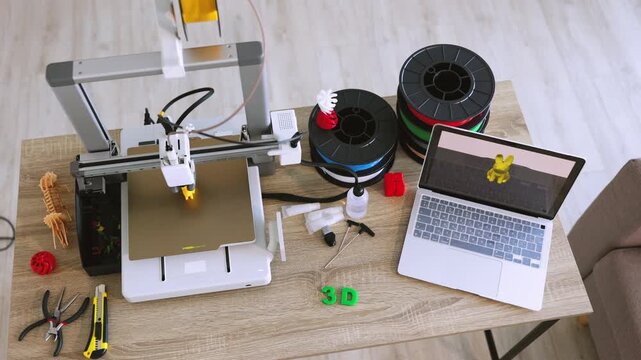 A 3D printer is actively creating a small yellow object while filament spools and tools are neatly arranged on a wooden desk next to a laptop displaying the printing process