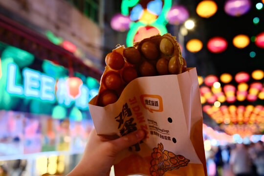 Hong Kong, HK, 10.03.2026: Hand holding street food takoyaki or egg waffle snack at colorful night market with lanterns