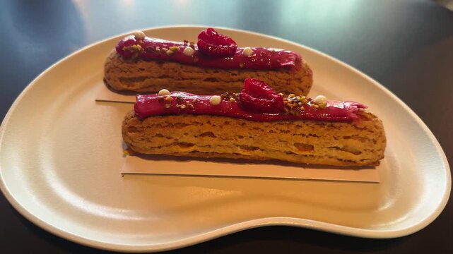 Fresh and delicious eclairs with sweet and sour raspberry cream, served on a white plate in a cozy French cafe. High quality 4k footage