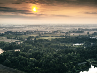 Malowniczy zachód słońca nad polskimi polami i lasami. Widok z góry na krajobraz rolniczy, wieś w oddali i nastrojową mgiełkę. Fotografia z drona. © Henryk Niestrój