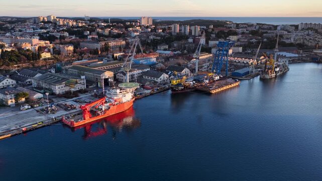Aerial view of a shipyard with a vibrant red vessel docked, juxtaposed against the city's buildings and the deep blue waters, Pula, Istria County, Croatia.