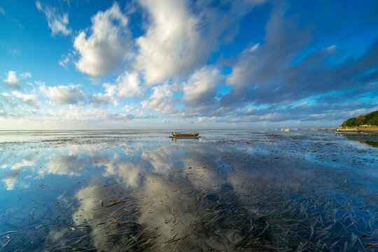 ast morning landscape at Sanur Beach, Bali featuring a tiny traditional boat reflecting in the shallow water against a massive dramatic sky