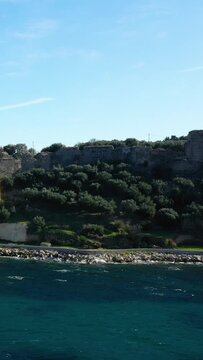 Scenic view of the turquoise sea along a rocky shoreline with lush greenery and an old stone wall in Koroni, Greece. Clear blue sky enhances the tranquil Mediterranean atmosphere.