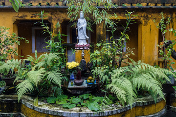 A serene Quan Am statue stands amidst lush greenery in Hoi An, Vietnam. The statue is adorned with offerings, creating a peaceful and spiritual atmosphere. © Zenstratus