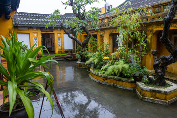 Obraz premium Rain falls on the courtyard of an Old House, a popular tourist attraction in the historic city of Hoi An, Vietnam. Lush plants thrive in the wet conditions.