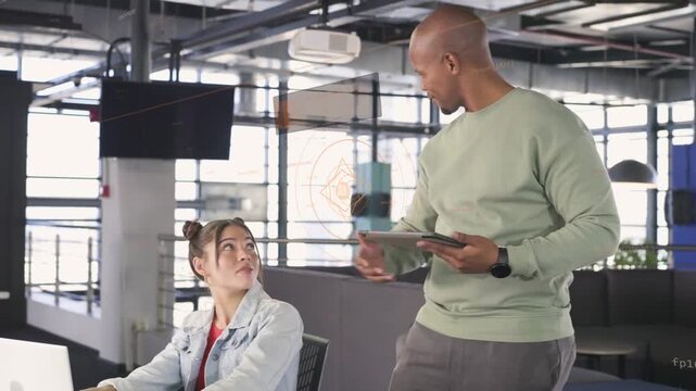 Male lead holding tablet and approaching female coworker in office, pointing at laptop, clarifying