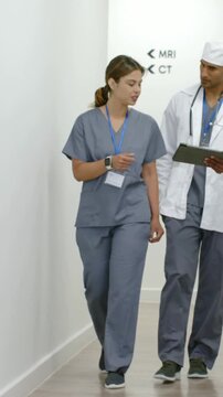 Vertical video: Walking scrub nurse and MD in coat holding tablet checking scan data by MRI CT sign