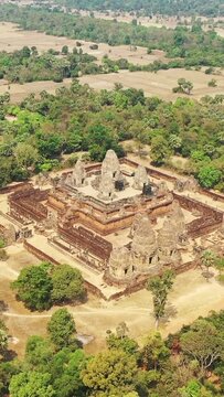 Drone shot of the ancient Pre Rup temple complex surrounded by trees and dry fields in Cambodia. The historic structure stands out with its unique architecture and natural surroundings.