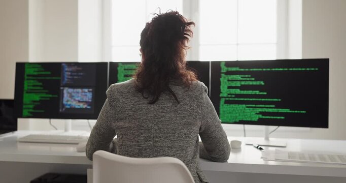 Developer programmer woman coding software on screens. Back view at multi monitor computer in bright office, coder typing on clean desk, focused on technology. Smart tech coding concept today.