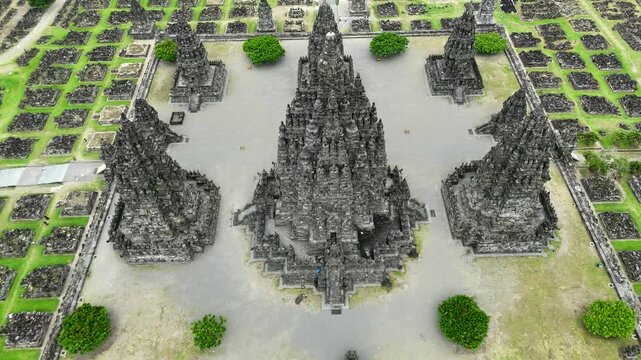 Cinematic aerial shot starting close to the stone spires of Prambanan Temple and smoothly pulling away to reveal the vast archaeological site and the surrounding rural landscape of Central Java under 