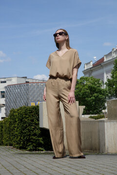 Fashionable Woman Walking in European City Street Wearing Monochrome Beige Flowy Trousers and V-Neck Short-Sleeve Shirt &ndash; Elegant Summer Style and Urban Lifestyle Concept