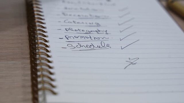 Close-up of a handwritten event planning checklist in a spiral notebook on a wooden desk, showing completed tasks like catering, photography, promotion, and schedule in soft indoor light.