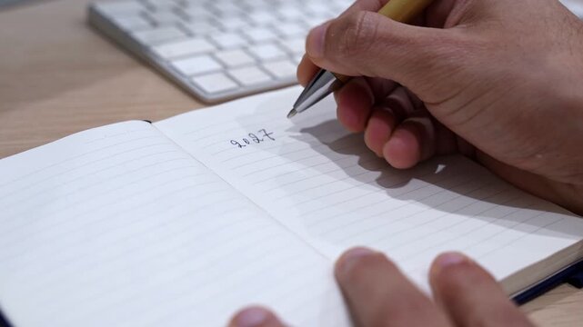 Close-up of a hand writing 2027 goals in an open notebook on a wooden office desk with pen and blurred keyboard, showing business and personal planning in an indoor workspace.