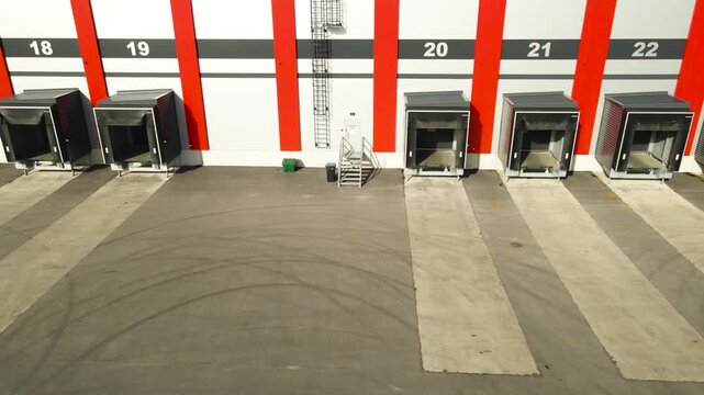 Low drone glide past numbered loading docks at a modern distribution center with empty bays bright sun and tire marked asphalt.