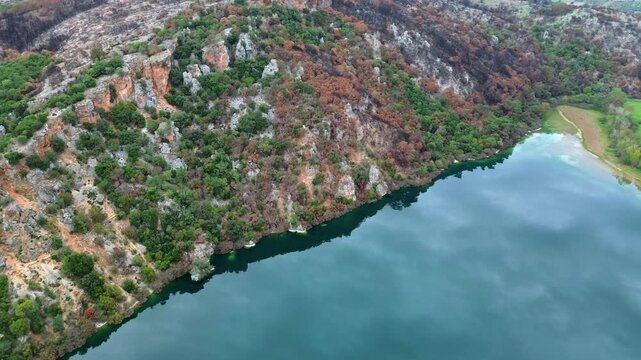 Cinematic drone shot of Lake Ziros in Preveza. High-angle view of rugged cliffs reflecting on the deep blue water, showing signs of past forest fires. Ideal for nature and environmental projects.